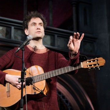 Man playing guitar speaking into mic in red jumper