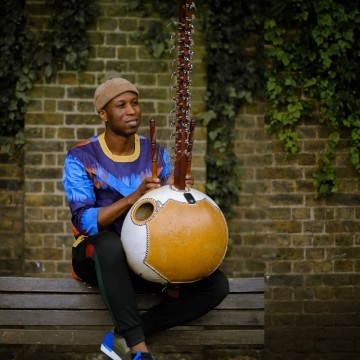 Man sitting on bench in multicolour top playing Kora instrument