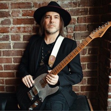 Man against brick wall wearing black hat holding guitar
