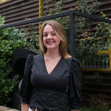 Blonde woman, black top standing in front on green bush